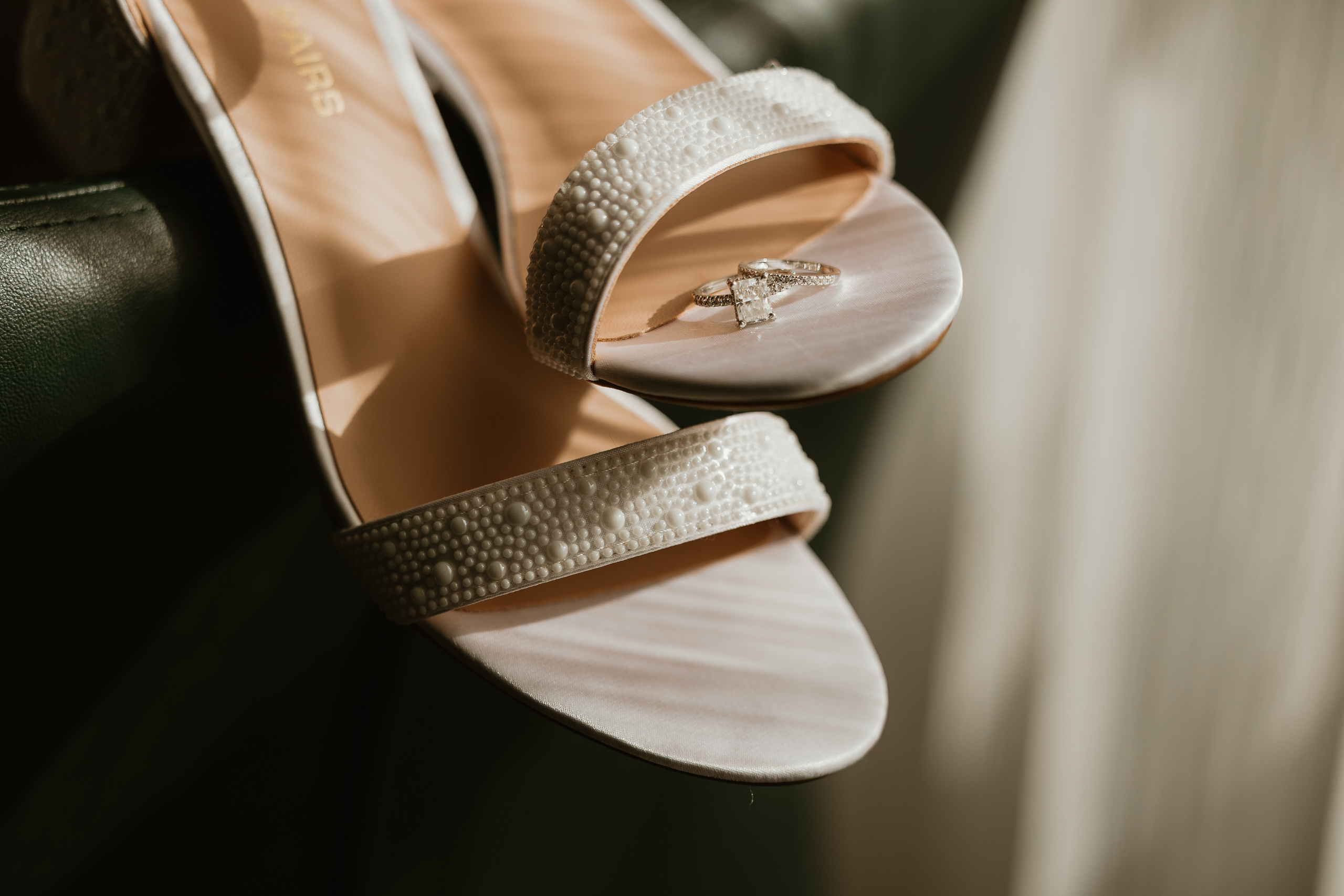 A pearl necklace elegantly laid next to a wooden ring box with a square-cut engagement ring and a pair of beaded bridal heels.